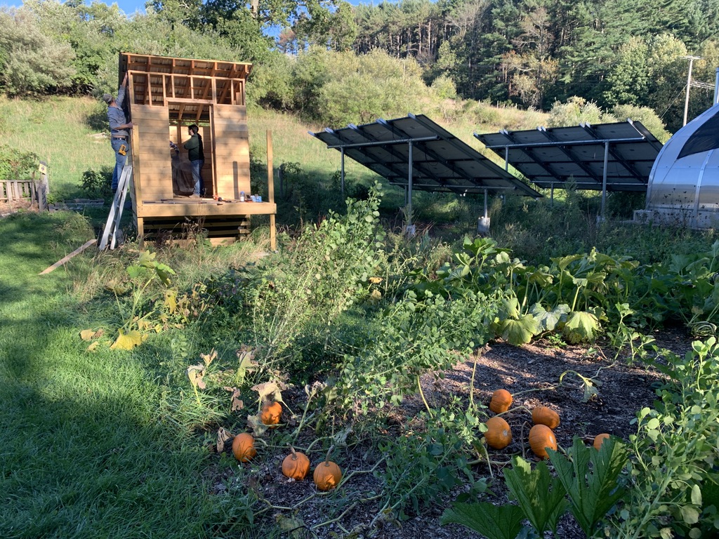 Volunteers build an outhouse against a grassy hillside, near some solar panels and a pumpkin patch.
